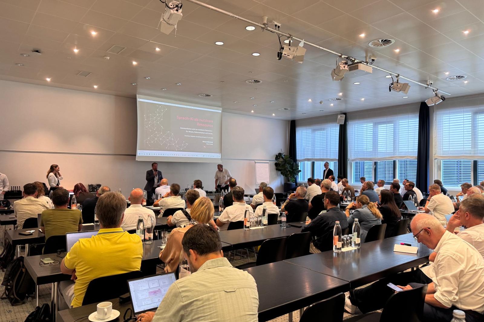 Voll besetzter Saal an der OneVoice-Tagung der Digitalen Verwaltung Schweiz, vorne die Titelfolie «Sprach-KI als nutzbare Ressource»